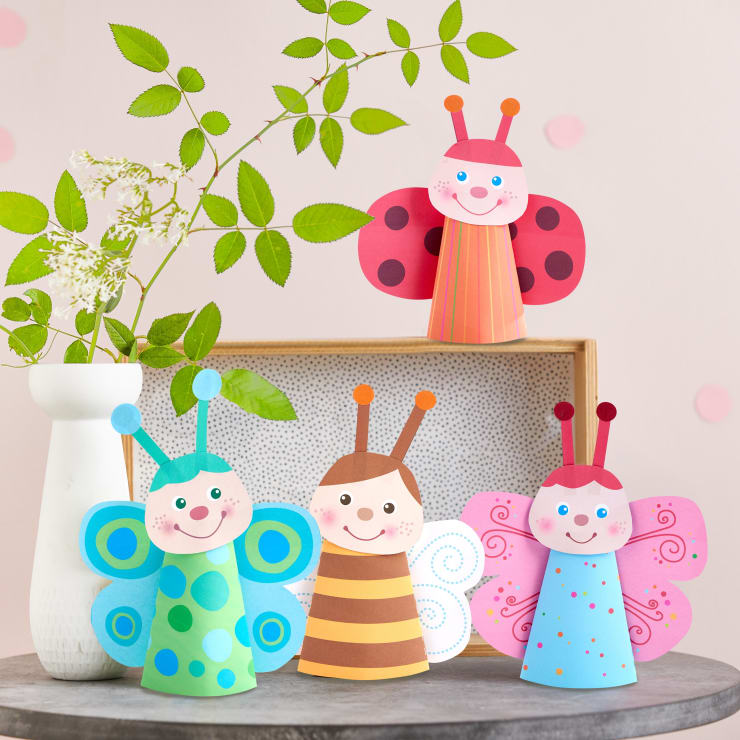 Vier bunte Papierkegel-Insekten, darunter ein Marienkäfer und drei Schmetterlinge, sind auf und vor einer Schachtel ausgestellt, mit einer weißen Vase mit grünem und weißem Laub auf der linken Seite vor einem hellrosa Hintergrund.