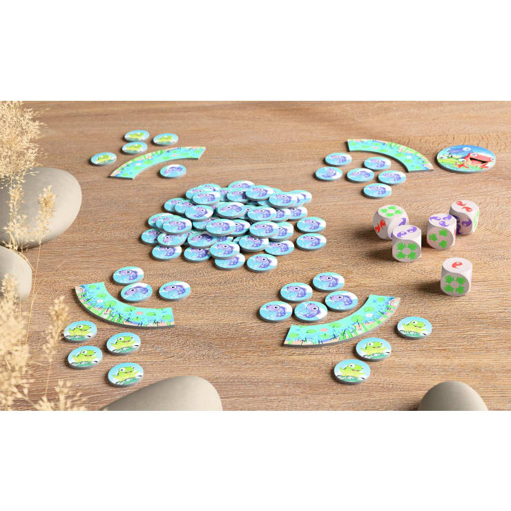 Ein Tischspiel mit blauen Froschplättchen, grünen Plättchen, vier Würfeln mit farbigen Symbolen und mehreren gebogenen Spielbrettern mit Cartoon-Fröschen, die alle auf einer Holzfläche angeordnet sind. Steine und trockene Pflanzen umrahmen die Szene.