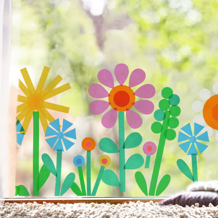 Bunte Papierausschnitte von verschiedenen Blumen und Blättern sind an einem Fenster mit einem verschwommenen grünen Hintergrund im Freien angebracht, der durch das Glas sichtbar ist. Die Blumenmuster sind geometrisch mit hellen, kräftigen Farben.