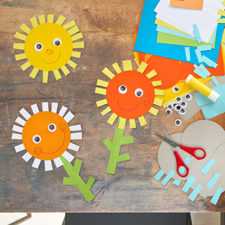 Drei Sonnenblumen aus Papier mit Kulleraugen und lächelnden Gesichtern sind auf einer Holzfläche angeordnet. Bastelmaterial wie farbiges Papier, Schere, Klebestift und Papierschnipsel liegen in der Nähe verstreut.