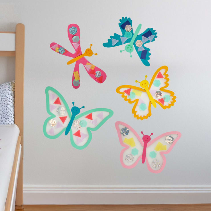 Bunte Papiermosaikschmetterlinge und eine Libelle sind an einer hellen Wand neben einem hölzernen Bettgestell ausgestellt. Die Insekten sind aus verschiedenen Formen und Farben von Papier hergestellt.