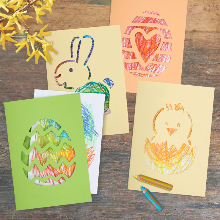 Vier farbige Papierkarten mit Ausschnitten zum Thema Ostern - ein Hase, ein Küken und zwei Eier -, die jeweils mit Buntstiftkritzeleien unterlegt sind. Zwei Buntstifte liegen nebeneinander auf einer hölzernen Unterlage, mit gelben Blumen in der oberen linken Ecke.