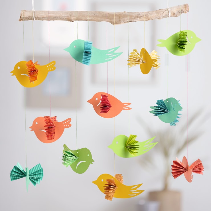 Ein Mobile aus einem Holzstab und bunten Papiervögeln, wobei jeder Vogel an einer Schnur hängt. Die Vögel sind in verschiedenen Farben und haben gefaltete Papierflügel, mit einem verschwommenen Hintergrund.