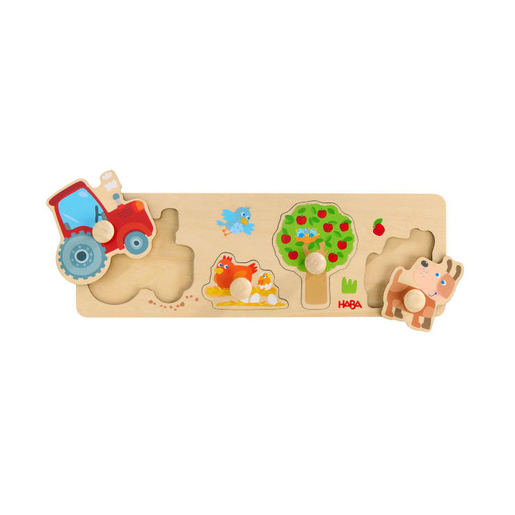 Ein Holzpuzzle mit drei Teilen: ein roter Traktor, ein grüner Baum mit Äpfeln und eine braune Kuh. Auf dem Puzzlebrett sind außerdem ein blauer Vogel und Heu abgebildet.