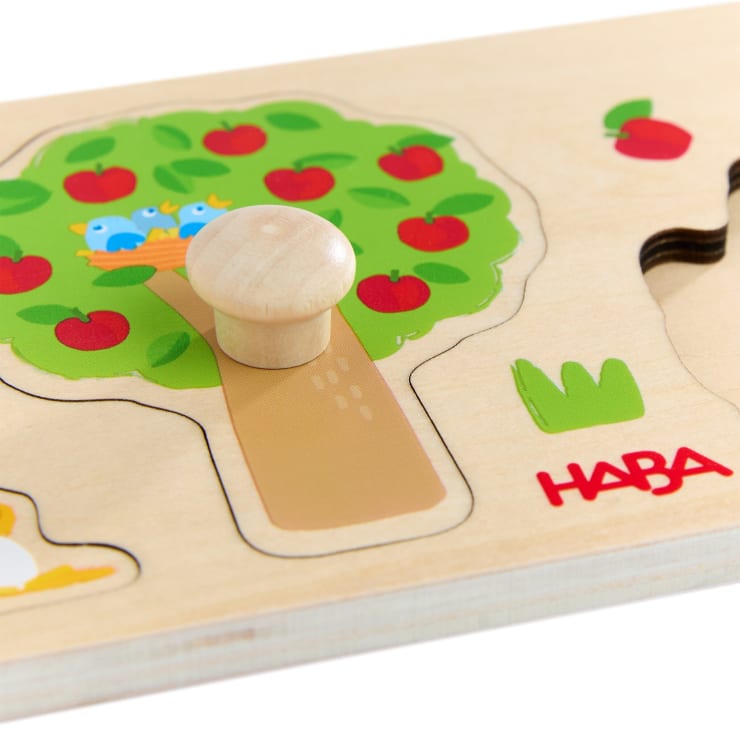 Nahaufnahme eines hölzernen Puzzleteils mit einem Knauf, auf dem ein Apfelbaum mit roten Äpfeln und zwei blauen Vögeln abgebildet ist. Das Puzzle ist mit dem roten HABA-Logo gebrandet.