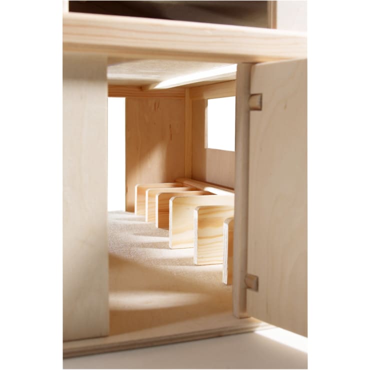Nahaufnahme eines kleinformatigen Architekturmodells aus Holz, das einen Raum mit vier hölzernen bankähnlichen Strukturen und einem Fenster an der rechten Wand zeigt. Die Tür auf der rechten Seite ist teilweise geöffnet.