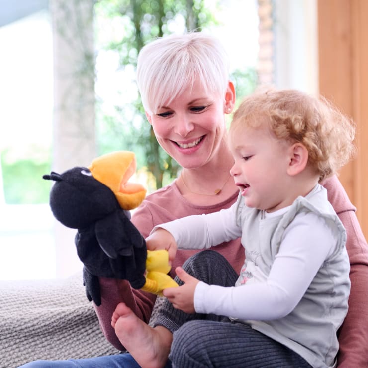 Ein Erwachsener mit kurzen weißen Haaren und ein kleines Kind sitzen zusammen auf einer Couch. Der Erwachsene hält eine schwarze Vogelhandpuppe, und das Kind interagiert mit ihr. Beide scheinen engagiert und auf die Puppe konzentriert zu sein.