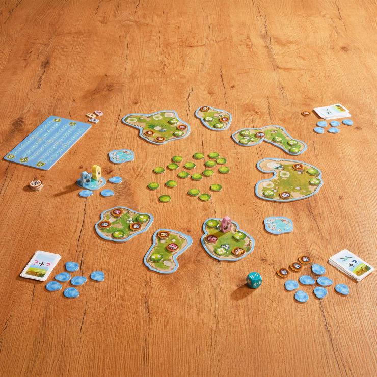 Auf einer hölzernen Fläche wird ein Brettspiel mit inselförmigen Spielsteinen, grünen und blauen Chips, Figuren, Würfeln, Karten und Punktetafeln aufgebaut, die im Kreis angeordnet sind.
