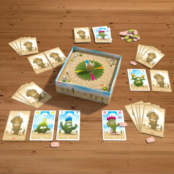 Auf einem Holztisch ist ein Brettspiel aufgebaut. Es besteht aus Cartoon-Kaktuskarten, einem runden Spielbrett in der Mitte mit einer grünen Drehscheibe, rosa und weißen Spielsteinen und zwei rosa Würfeln. Mehrere Kartenblätter sind um das Spielbrett herum angeordnet.