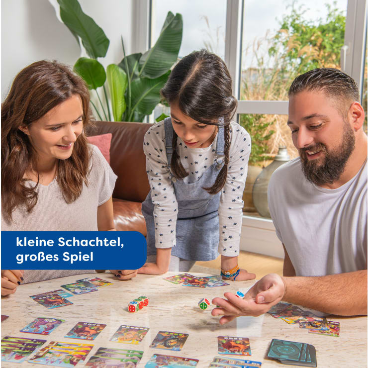 Drei Personen, zwei Erwachsene und ein Kind, sitzen an einem Tisch und spielen ein buntes Karten- und Würfelspiel in einem hellen Wohnzimmer. Der Text auf dem Bild lautet: "Kleine Schachtel, großes Spiel".