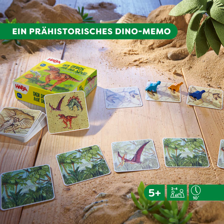 Ein Dinosaurier-Memory-Kartenspiel wird auf einer sandigen Fläche mit vier kleinen Dinosaurierfiguren und mehreren illustrierten Karten aufgebaut. Die Spielschachtel und die Anleitung sind sichtbar. Die Symbole zeigen an, dass das Spiel für 2 bis 4 Spieler ab 5 Jahren geeignet ist und 10 Minuten dauert.