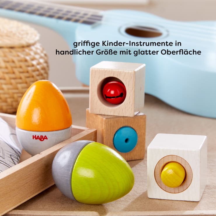 Eine blaue Ukulele, ein Holzkorb, eiförmige Maracas und Holzklötze mit rotierenden farbigen Zentren sind auf einer hellen Fläche angeordnet. Der deutsche Text oben beschreibt kindgerechte Instrumente mit glatten Oberflächen.