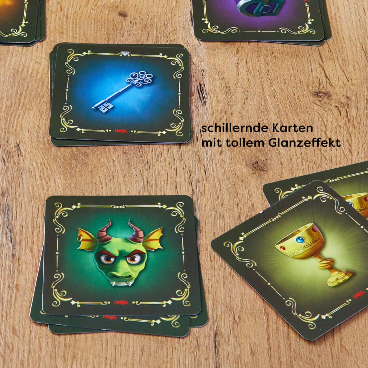 Auf einem Holztisch liegen mehrere illustrierte Spielkarten mit verschnörkelten Rändern. Die Karten zeigen Bilder wie einen Schlüssel, einen Drachenkopf und einen goldenen Becher. Der deutsche Text beschreibt den schimmernden Glanzeffekt der Karten.
