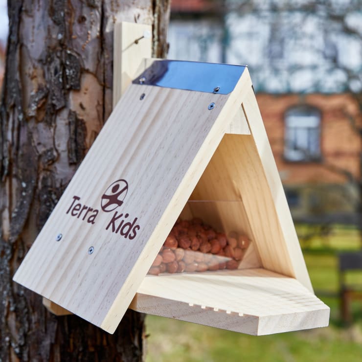 Ein dreieckiges Vogelfutterhaus aus Holz mit der Aufschrift Terra Kids ist an einem Baumstamm befestigt. Das Futterhaus hat ein Metalldach und enthält im Inneren braune Nüsse oder Samen. Im Hintergrund sind ein verschwommenes Gebäude und Bäume zu sehen.