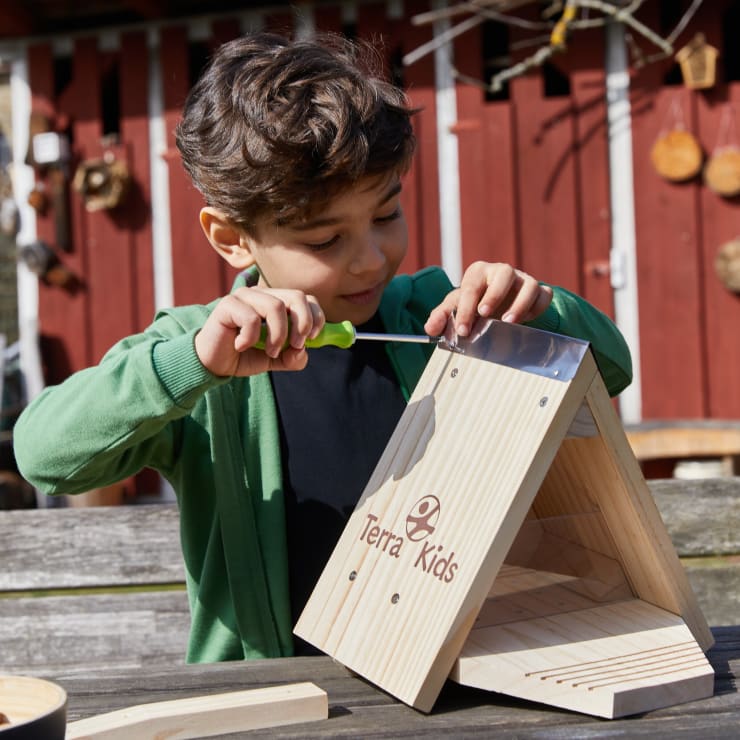Ein Junge sitzt draußen an einem Holztisch und setzt mit einem Schraubenzieher ein hölzernes Terra Kids Projekt zusammen. Im Hintergrund ist eine rote Scheune mit hängenden Dekorationen zu sehen.