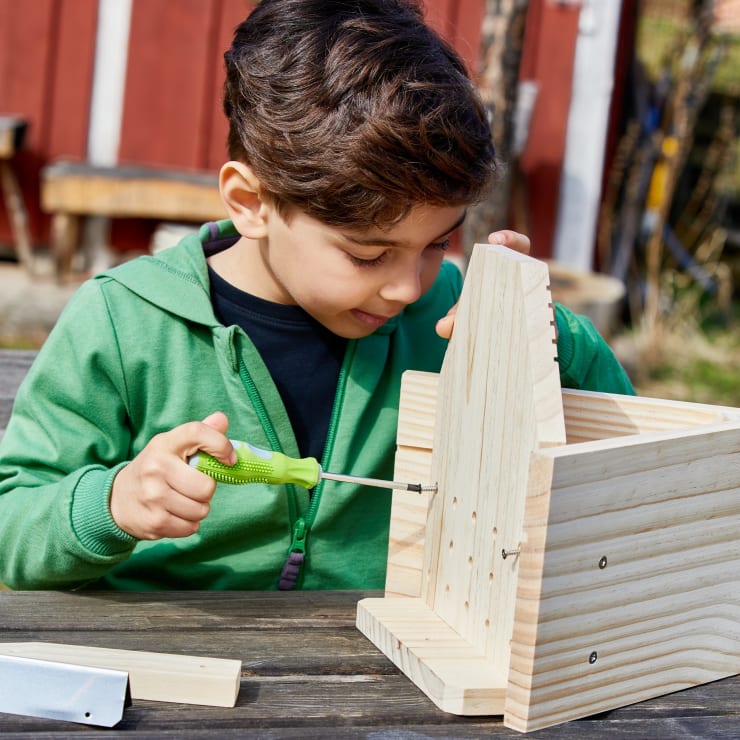 Ein Junge in einer grünen Jacke setzt mit einem Schraubenzieher eine Holzkonstruktion an einem Tisch im Freien zusammen. Neben ihm liegen Holzstücke und Werkzeuge, und im Hintergrund ist ein rotes Gebäude zu sehen.