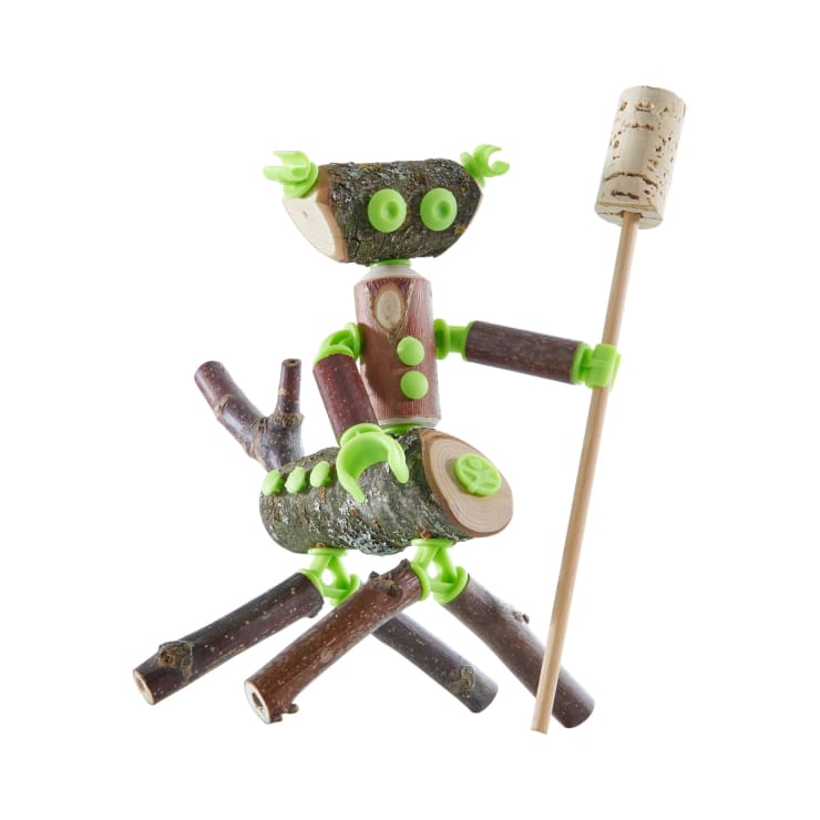 Eine kleine Figur aus verschiedenen Stöcken und Holzteilen, mit grünen Plastikverbindern, hält einen Stock mit einem Korken an der Spitze vor einem weißen Hintergrund.