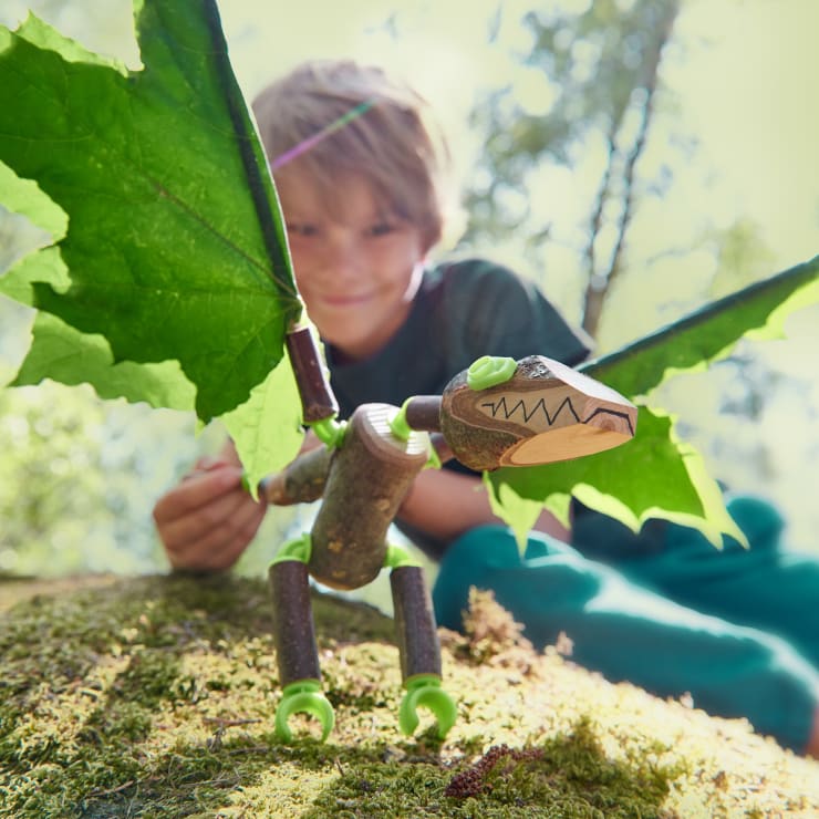 Ein Kind hockt im Freien hinter einer handgefertigten Drachenfigur aus Stöcken, Blättern und Spielzeuggelenken, die auf moosigem Boden steht, während das Sonnenlicht durch die Bäume im Hintergrund fällt.