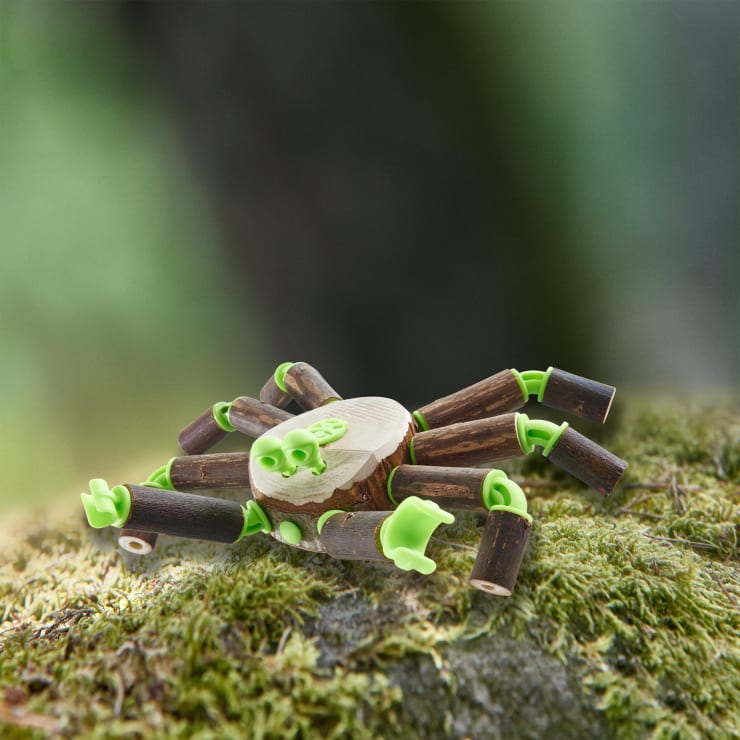 Eine kleine Spielzeugspinne aus grünen Plastiksteckern und braunen Holzstäben sitzt auf Moos, wobei der unscharfe grüne Hintergrund eine natürliche Umgebung im Freien andeutet.