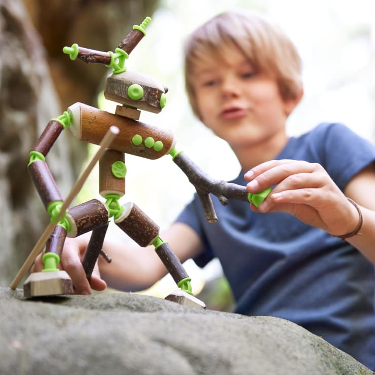 Ein Kind in einem blauen Hemd spielt mit einer handgefertigten Roboterfigur aus Holz, die aus Stöcken und grünen Plastikverbindern zusammengesetzt ist, im Freien auf einem großen Felsen.