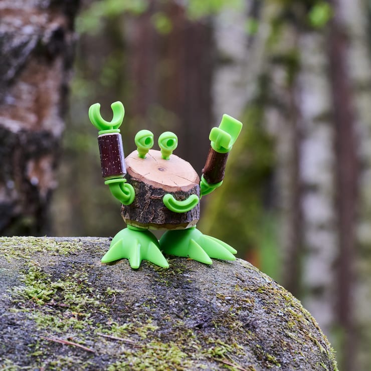 Ein kleines Spielzeugwesen mit einem hölzernen Körper, grünen Plastikbeinen und -krallen und hervorstehenden Augen steht auf einem moosbewachsenen Felsen in einer Waldlandschaft.
