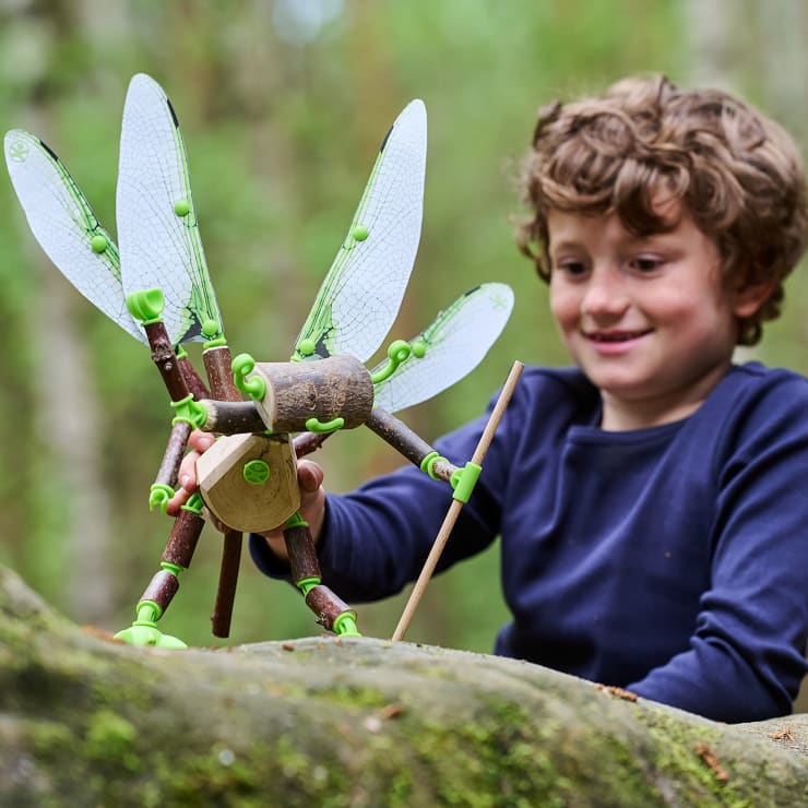 Ein kleiner Junge mit lockigem Haar lächelt, während er im Freien mit einem großen Stabheuschreckenmodell mit durchsichtigen Flügeln spielt, das auf einem moosbewachsenen Felsen in einem Wald aufgebaut ist.