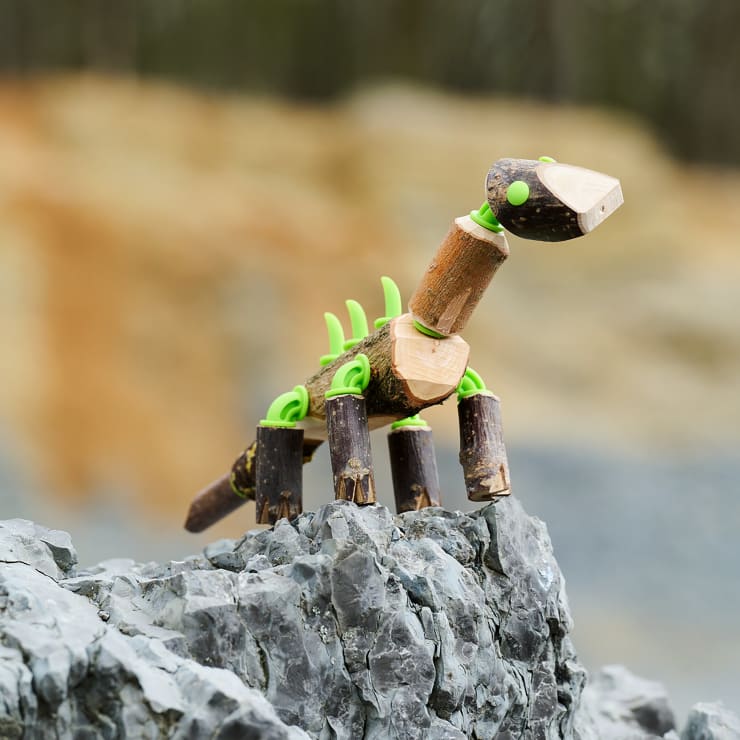 Eine kleine Dinosaurierfigur aus Holzstäben und grünen Plastikteilen steht auf einer grauen, felsigen Oberfläche im Freien, mit einem unscharfen Hintergrund aus Steinen und Erde.