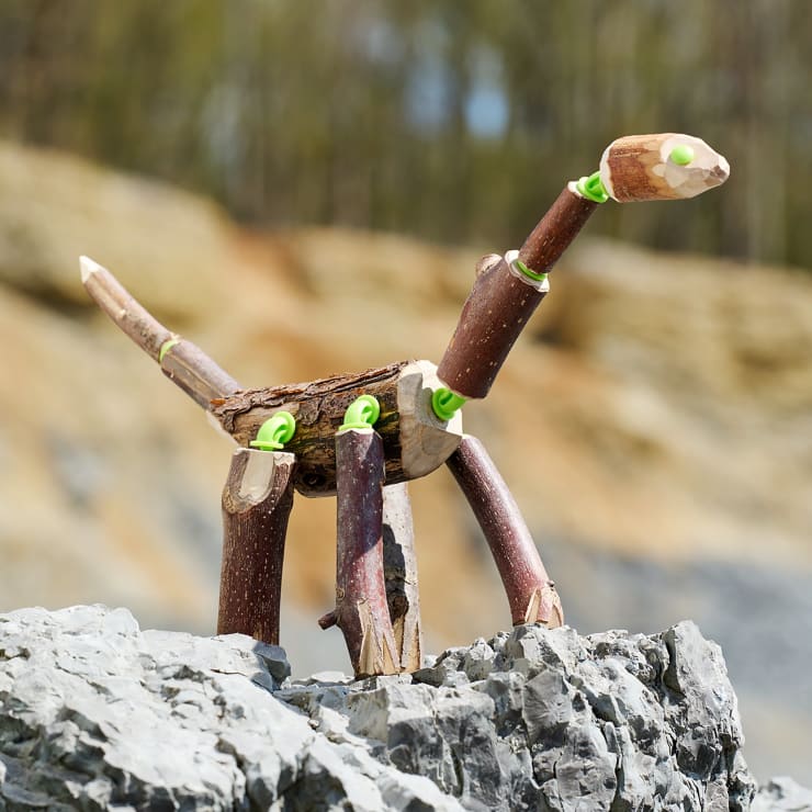 Eine kleine Dinosaurierfigur aus Stöcken, Rinde und grünen Steckverbindungen steht auf einem grauen Felsen, mit einem unscharfen Hintergrund im Freien.
