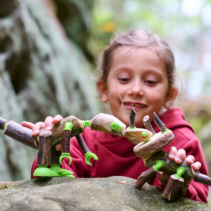 Ein junges Mädchen mit rotem Kapuzenpulli spielt im Freien mit Spielzeugdinosauriern aus Naturholz und grünen Plastikteilen und stellt sie auf einen großen Felsen mit unscharfem Naturhintergrund.