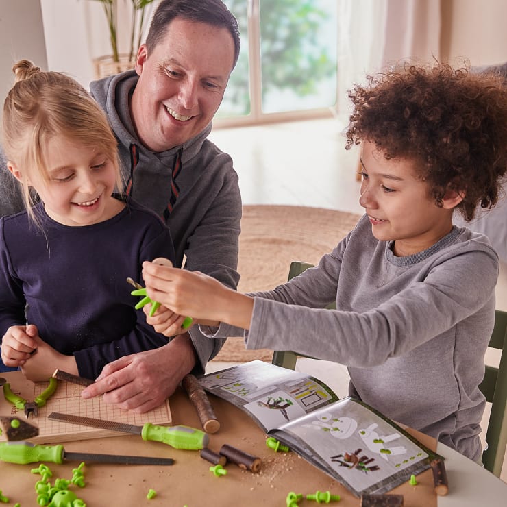 Ein Erwachsener und zwei Kinder sitzen an einem Tisch und bauen ein Modell aus grünen Plastikteilen zusammen, wobei sie Werkzeuge benutzen und den Anweisungen in einem aufgeschlagenen Heft folgen. Sie lächeln und arbeiten zusammen in einem hell erleuchteten Raum.