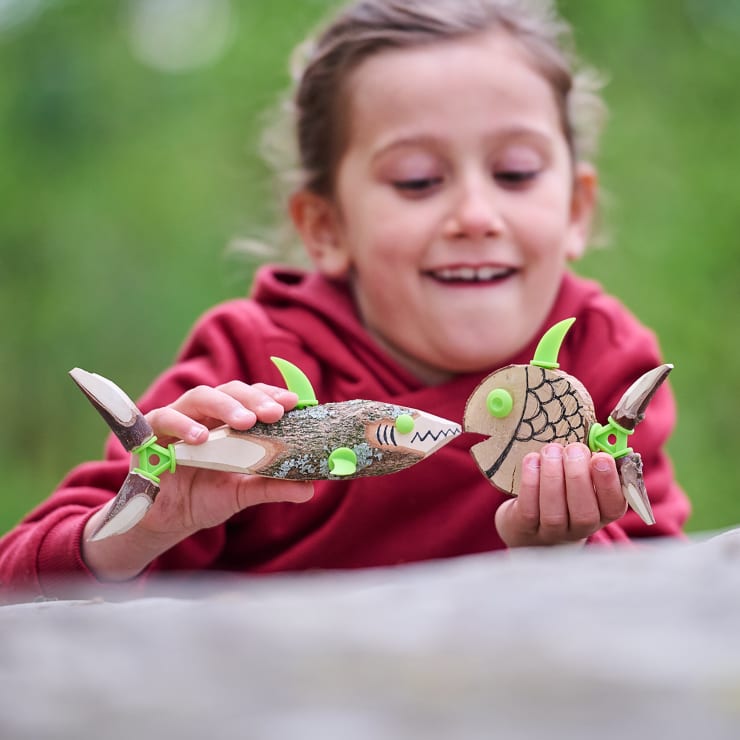 Ein Kind in einem roten Kapuzenpulli lächelt, während es zwei Spielzeugfische aus natürlichen Materialien wie Holz und Blättern mit grünen Kunststoffteilen in der Hand hält. Der Hintergrund besteht aus unscharfem Grün.