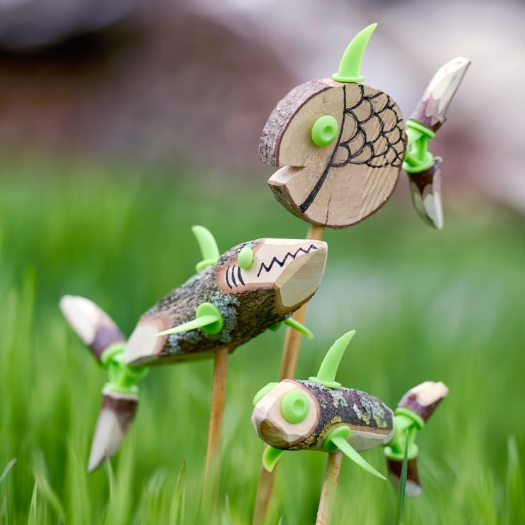 Drei handgefertigte Fische aus Holz und grünen Kunststoffteilen sind auf Stöcken montiert und aufrecht im grünen Gras aufgestellt. Die Fische sind mit einfachen schwarzen Mustern versehen, die Schuppen und Gesichtszüge darstellen.