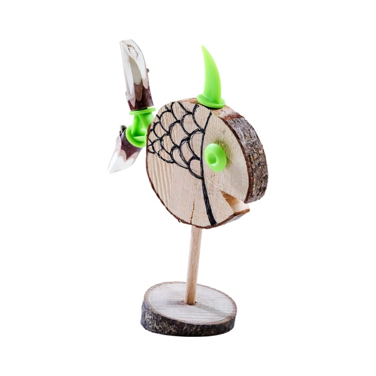Eine hölzerne Fischfigur mit einfachen schwarzen Linien, einer grünen Plastikflosse und einem Auge, montiert auf einem hölzernen Ständer, vor einem weißen Hintergrund.