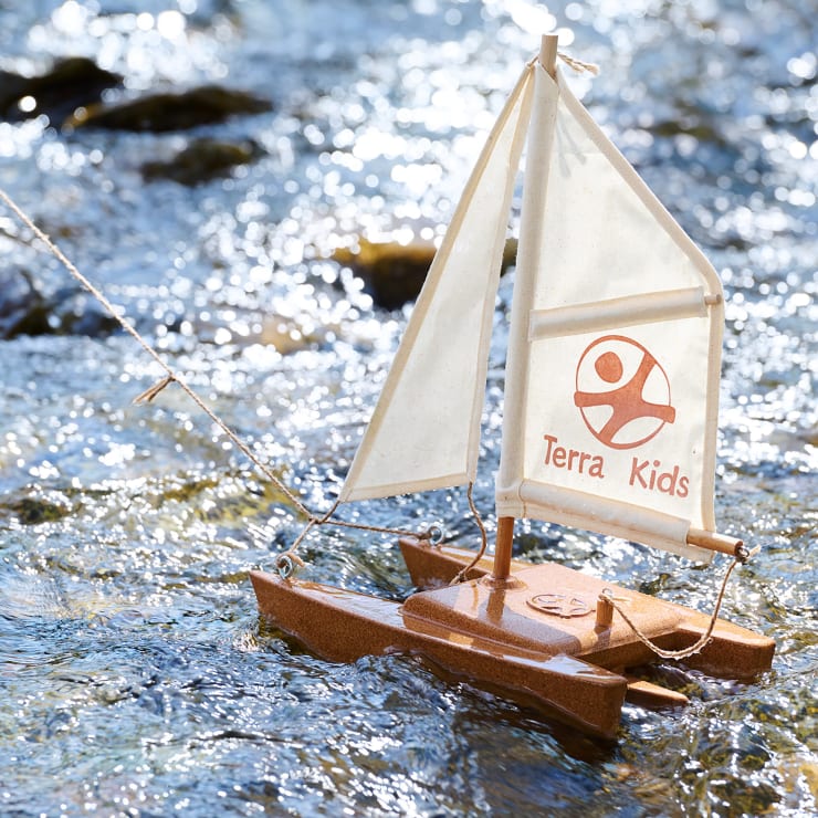 Ein kleines Modell eines Segelboots mit weißen Segeln und dem Aufdruck Terra Kids schwimmt an einer Schnur befestigt auf dem Wasser. Das Sonnenlicht spiegelt sich auf der kräuselnden Oberfläche um das Boot herum.
