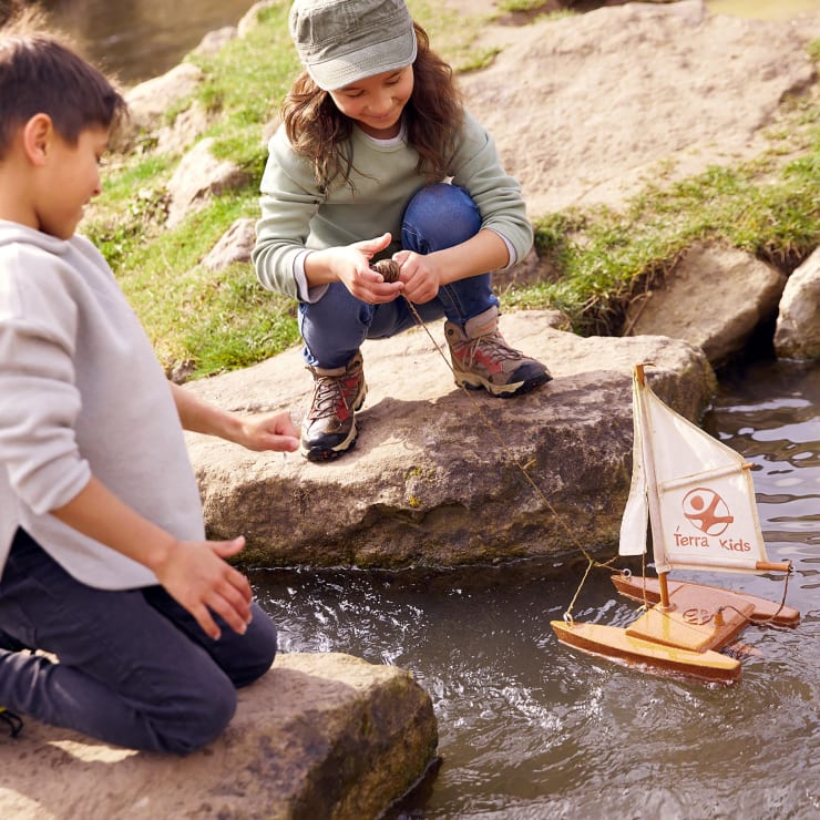 Zwei Kinder spielen an einem Bach und sitzen auf Steinen. Ein Kind hält einen Zweig, während das andere ein kleines hölzernes Segelboot mit der Aufschrift Terra Kids im Wasser lenkt. Um sie herum sind Gras und Felsen zu sehen.