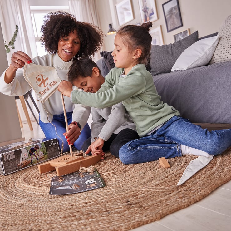 Ein Erwachsener und zwei Kinder sitzen auf einem gewebten Teppich in einem Wohnzimmer und bauen einen Bausatz für ein Holzspielzeugboot zusammen. Der Erwachsene hält eine Stofftasche mit der Aufschrift "Terra Kids" in der Hand, während die Kinder gemeinsam an den Bootsteilen arbeiten.