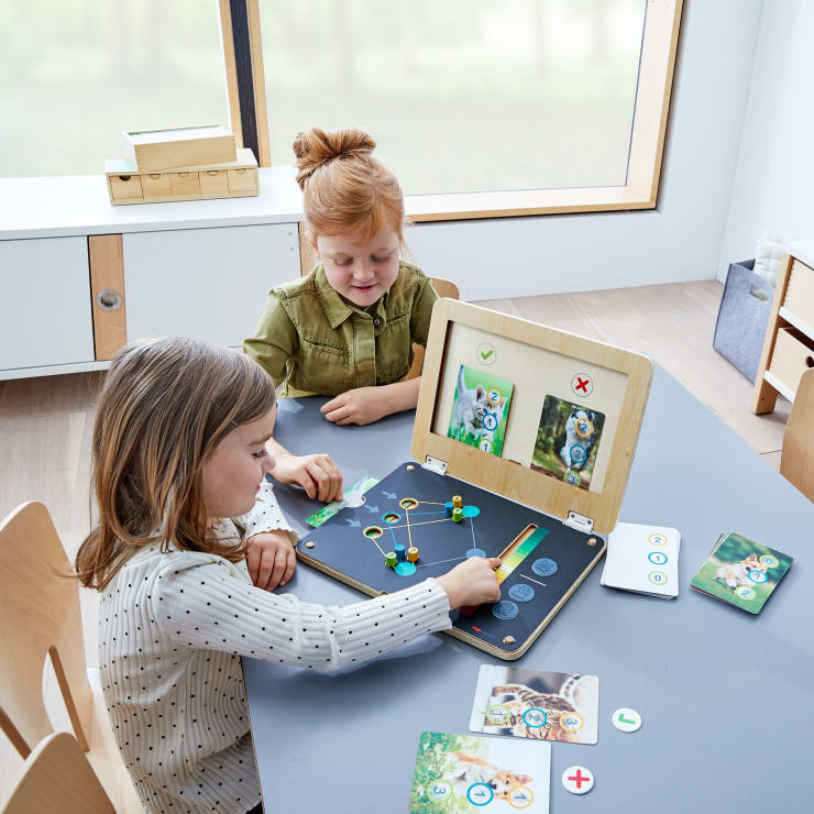 Zwei kleine Kinder sitzen an einem Tisch und spielen ein pädagogisches Brettspiel mit Tierkarten, Spielsteinen und einem hölzernen Spielbrett. Im Hintergrund sind ein großes Fenster und Regale zu sehen.