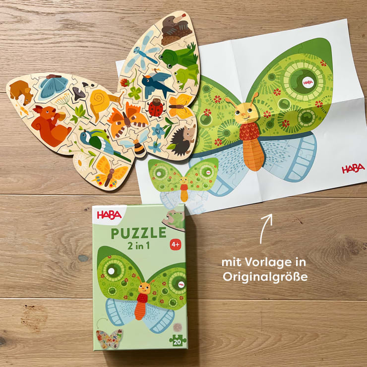 Ein hölzernes Schmetterlingspuzzle mit Tierillustrationen, ein Referenzblatt aus Papier, das denselben Schmetterling zeigt, und eine grüne HABA 2-in-1-Puzzlebox liegen auf einer Holzfläche. Der Text auf Deutsch befindet sich auf dem Bild.