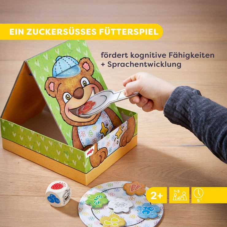 Eine Kinderhand füttert mit einer Pappzange einen lächelnden Bären, der in einer dreieckigen Schachtel abgebildet ist. Auf dem Tisch befinden sich Würfel und ein Teller mit Essensmarken. Der deutsche Text beschreibt das Spiel als Förderung der kognitiven und sprachlichen Fähigkeiten.
