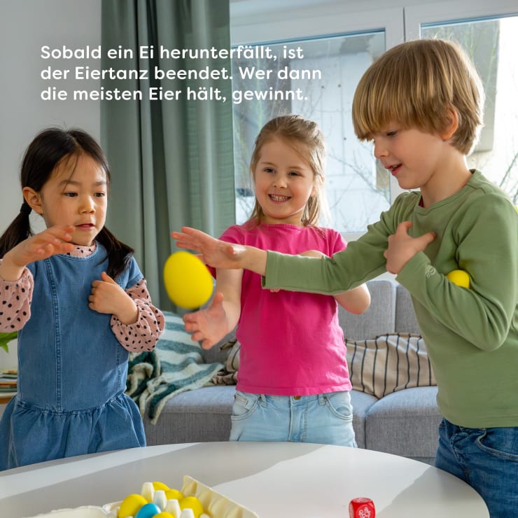 Drei kleine Kinder stehen in einem Raum und balancieren sorgfältig ein gelbes Plastikei auf einem Löffel. Vor ihnen befindet sich ein Tisch mit weiteren gelben Eiern und Eierbechern. In der oberen linken Ecke ist ein weißer Text in deutscher Sprache zu lesen.