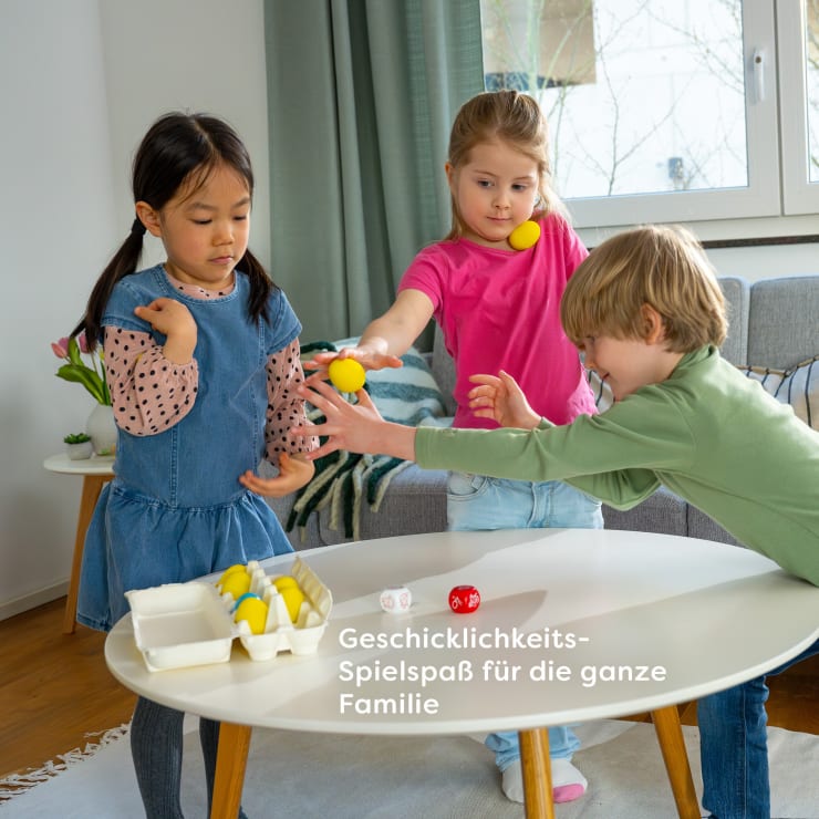 Drei Kinder spielen in einem hellen Wohnzimmer ein Tischspiel mit gelben Bällen. Ein Kind streckt die Hand aus, während ein anderes einen Ball hält und das dritte auf seinen Arm schaut. Der Text auf dem Tisch lautet: Geschicklichkeits-Spielspaß für die ganze Familie.