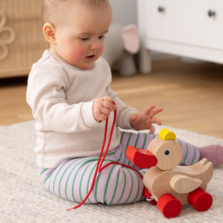 Ein Baby sitzt auf einem hellen Teppich und hält eine rote Schnur in der Hand, an der ein hölzernes Enten-Nachziehspielzeug befestigt ist. Das Kind trägt ein beiges Oberteil und eine gestreifte Hose. Im Hintergrund sind ein Schrank und ein Plüschtier zu sehen.