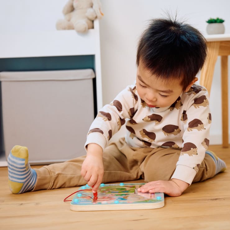 Ein kleines Kind sitzt auf einem Holzboden und spielt mit einer magnetischen Labyrinth-Tafel. Das Kind trägt ein igelgemustertes Hemd, eine hellbraune Hose und unpassende Socken. Im Hintergrund sind ein Kuscheltier und ein Vorratsbehälter zu sehen.
