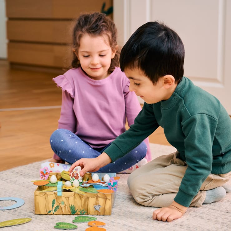 Ein junges Mädchen und ein Junge sitzen drinnen auf einem Teppich und spielen mit einem Brettspiel, das kleine Tier- und Eierfiguren enthält. Der Junge versucht, eine Figur zu bewegen, während das Mädchen lächelnd zusieht.