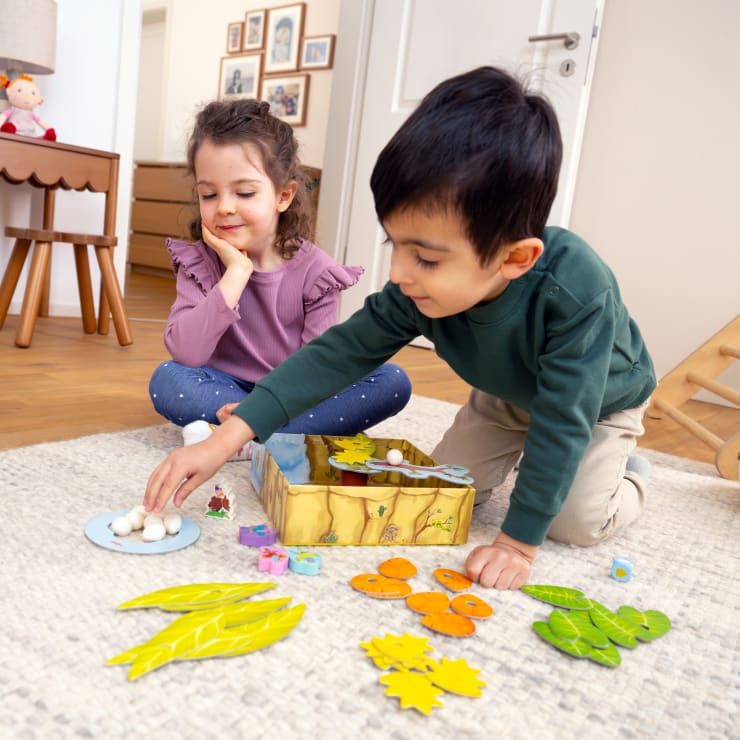Zwei kleine Kinder sitzen auf dem Boden und spielen ein Brettspiel mit Papierblättern, Eiern und Tierfiguren, die vor ihnen in einem hell erleuchteten Wohnzimmer ausgebreitet sind.
