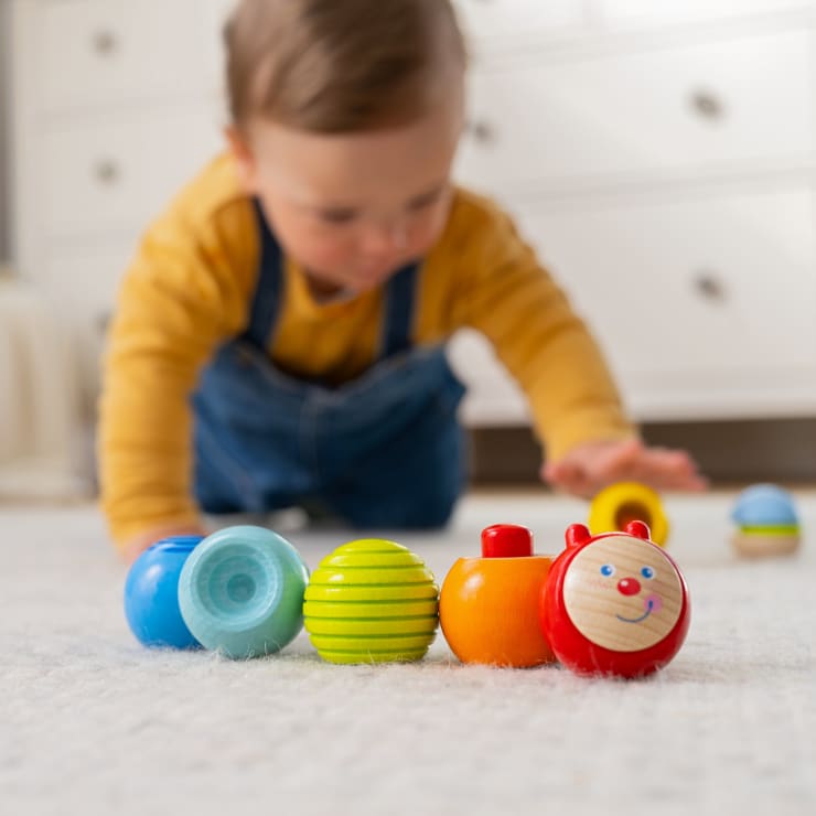 Ein kleines Kind in einem gelben Hemd und einem blauen Overall krabbelt auf einem Teppichboden und greift nach buntem Holzspielzeug, darunter ein rotes Spielzeug mit einem lächelnden Gesicht, vor einer verschwommenen Kommode im Hintergrund.