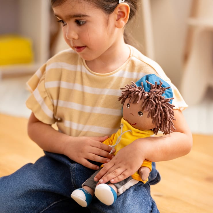 Ein kleines Kind mit dunklen Haaren sitzt auf dem Boden und hält eine Stoffpuppe, die mit einem gelben Kapuzenpulli, einer grauen Hose und einem blauen Hut bekleidet ist. Das Kind trägt ein gelb-weiß gestreiftes Hemd und eine blaue Hose.