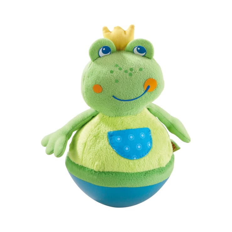 Ein grüner Plüschfrosch mit einer gelben Krone, orangefarbenen Wangen, einer blauen Tasche auf dem Bauch und einem abgerundeten blauen Boden, der aufrecht vor einem weißen Hintergrund steht.