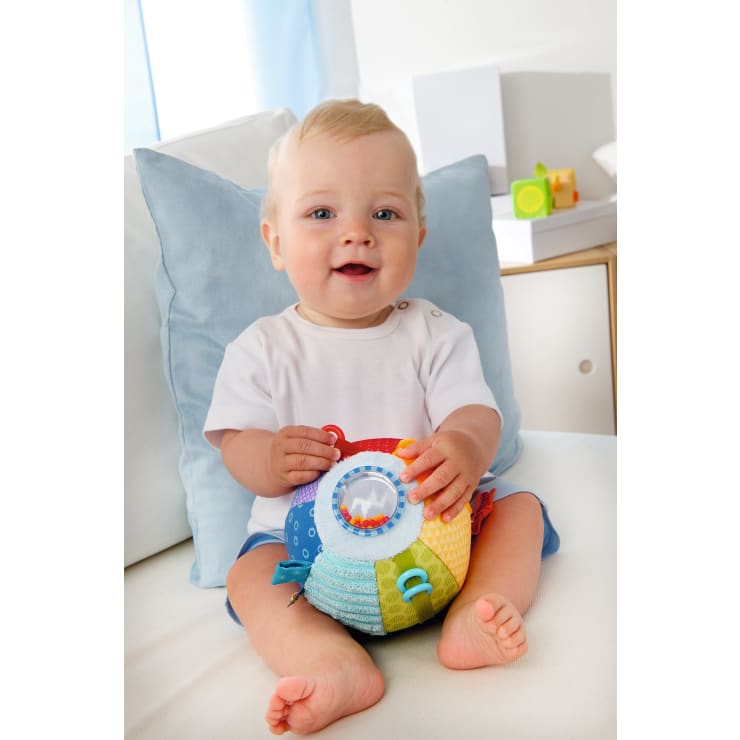 Ein Baby mit blondem Haar sitzt auf einer weißen Couch und hält ein buntes Sinnesspielzeug mit verschiedenen Strukturen und Aufsätzen, darunter ein durchsichtiger Teil mit Perlen. Hinter dem Baby befindet sich ein hellblaues Kissen.