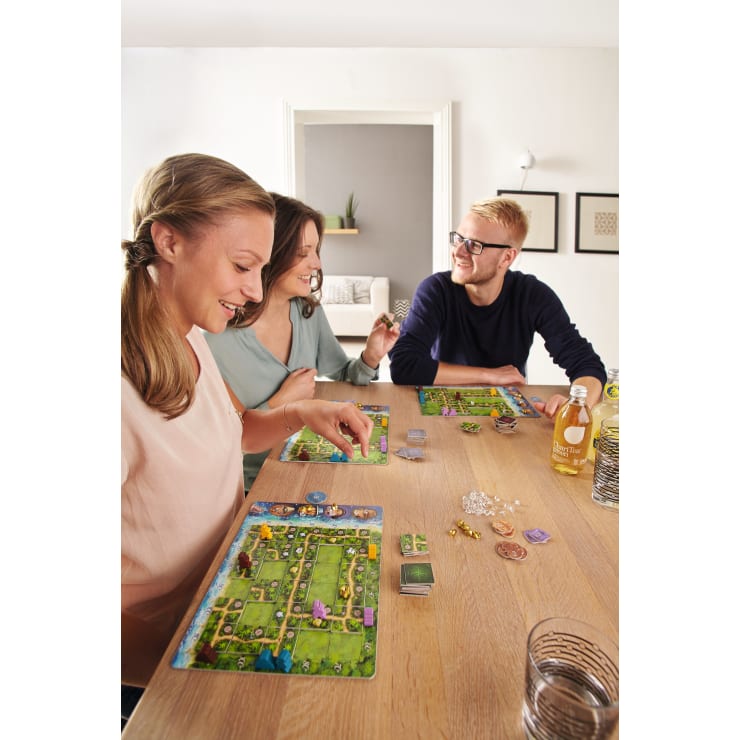 Drei Personen sitzen an einem Holztisch und spielen ein Brettspiel. Auf dem Tisch befinden sich Spielbretter, Karten, Spielsteine und Getränke. Im Hintergrund sind eine weiße Wand, ein Türrahmen und gerahmte Bilder zu sehen. Alle drei scheinen mit dem Spiel beschäftigt zu sein.